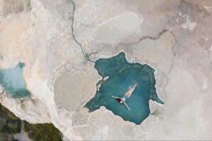 Aerial view of a person floating in a turquoise natural pool surrounded by textured beige rock formations.